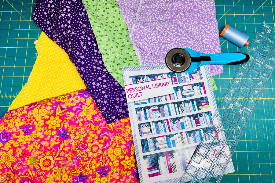 Materials in place to make the bookshelf quilt include fabric, pattern, rotary cutter, ruler, mat and thread.